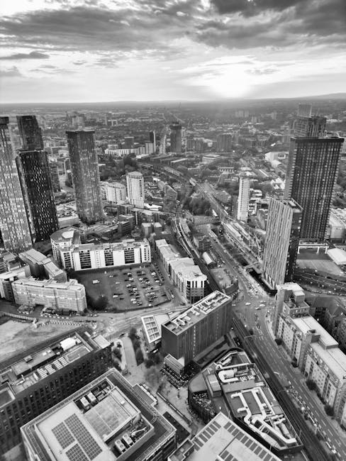 A high-angle black-and-white aerial photograph of a dense urban cityscape showing numerous modern high-rise office buildings and residential towers, with some mid-rise structures and parking lots visible. The scene captures busy streets, railway tracks, and a variety of construction materials scattered across a parking area, indicating ongoing building or moving activities. The sky above has scattered clouds, and the overall view illustrates a metropolitan area likely in White City, with the city’s skyline extending toward the horizon. The image, associated with Man with Van White City and their house removals services, highlights the importance of careful planning during home relocation or furniture transport within the city, emphasizing the busy urban environment involved in moving logistics.