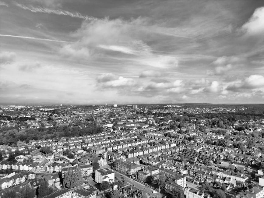 A black and white aerial photograph of a densely populated residential area in White City, showing rows of terraced houses with pitched roofs, streets lined with parked cars, and larger commercial or community buildings scattered throughout. The image captures the expanse of the urban landscape extending to the horizon under a sky filled with scattered clouds and wispy contrails. In the foreground, rooftops and street layouts are clearly visible, with some trees and greenery interspersed among the houses. The overall scene reflects a typical suburban neighbourhood, with the arrangement of homes and the urban density indicative of a property market focused on house relocations and removals, in alignment with services offered by Man with Van White City.