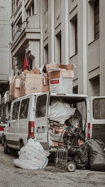 The image depicts the rear of a white cargo van parked on a city street in White City, with its back doors open revealing an interior filled with various packed materials including large plastic bags, cardboard boxes, and protective wrapping. Several cardboard boxes and packing materials are stacked on top of the van, some of which are labeled. A small hand trolley and protective blankets are visible nearby, indicating the ongoing loading process during a home relocation or furniture transport. The backdrop consists of multi-storey buildings with beige facades and several windows, suggesting an urban environment. The scene captures the typical logistical elements involved in professional removals, such as careful packing, loading, and transportation of household items, with the presence of company branding from Man with Van White City subtly informing the context of the moving service.