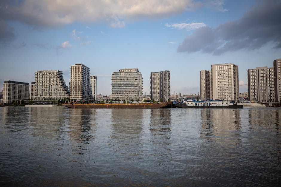 A panoramic view of modern high-rise residential buildings along a riverbank, with the buildings featuring a combination of glass windows, balconies, and varying architectural designs. The river in the foreground reflects the cloudy sky above, showing gentle ripples on its surface. The scene is set during daytime with natural light illuminating the skyline and water. This urban riverside landscape relates to house removals and property relocations in White City, with the buildings indicating a densely populated area suitable for professional moving services by Man with Van White City, as part of home relocation and furniture transport processes.