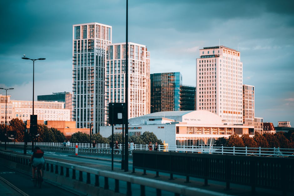 A cityscape of modern high-rise buildings illuminated by warm natural light, featuring a mix of tall glass and concrete structures with varying window patterns. In the foreground, there is a road with a cyclist riding along the pavement and a pedestrian walking nearby. Street lighting poles and traffic barriers are visible along the road. Below the buildings, a white, curved-roof building suggests an entertainment or conference venue, surrounded by trees and lower-rise structures. The scene appears to capture an urban area suitable for home relocation and furniture transport services by Man with Van White City, with the environment highlighting the busy yet organized atmosphere typical of city-based removals, where items are often moved through roads and loading areas near apartment complexes or office buildings.