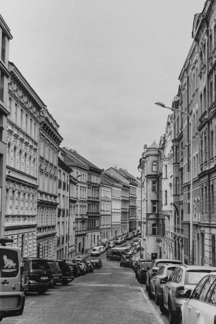 A narrow street in White City with tall, historic-style terraced buildings on both sides, featuring detailed facades and bay windows. Parked cars line both sides of the street, including hatchbacks, sedans, and vans, with some vehicles partially visible behind others. The pavement shows various cardboard boxes, wrapped furniture pieces with plastic or fabric covers, and moving supplies arranged near the edge of the sidewalk, indicating preparations for a home relocation or furniture transport. A man with a trolley or dolly is visible, actively loading or moving items from the building toward a nearby van parked at the curb. The scene is well-lit with natural daylight, and a streetlight extends from a lamppost on the right side. This image captures the logistics involved in a professional removal or moving service, with evidence of packing, furniture handling, and vehicle loading on a typical city street. Man with Van White City is engaged in the loading process, highlighting the house removal activity in this urban environment.
