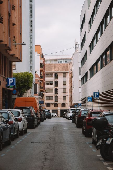 A narrow urban street scene in White City with parked cars lining both sides of the road, including compact hatchbacks, sedans, and a small van. The street is bordered by multi-storey residential and commercial buildings constructed with modern materials such as concrete, glass, and metal. Some buildings feature balconies, while others have large windows. A couple of parking signs indicating designated parking areas are visible, along with a street lamp mounted on a building on the left side. The sky is overcast, and the street appears clean and calm, likely during daytime. This setting illustrates a typical location for home relocation or furniture transport, with space for loading items onto service vehicles, supporting the relocation services offered by Man with Van White City, accommodating moving logistics in busy urban environments.