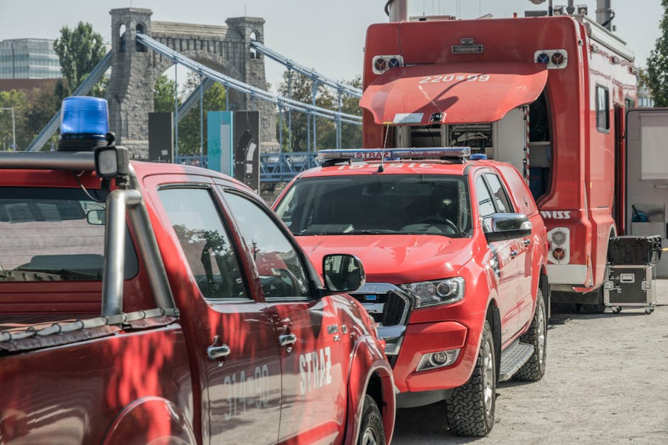 A scene during a home relocation or furniture transport process featuring multiple emergency service vehicles, including a red fire engine with its side panels open, and a red police car with a blue emergency light on top, parked on a paved surface. The fire engine's rear compartment contains equipment boxes, and the police vehicle is positioned adjacent to it. Behind the vehicles, a second presence of another emergency vehicle suggests a coordinated effort. In the background, there is a bridge with stone supports and suspended cables, along with modern office buildings and green trees, indicating an urban environment. The image captures a moment of loading or unloading activities, with no visible movement of furniture or boxes but emphasizing the vehicles used in large-scale or urgent removals, possibly as part of a logistical or safety protocol during a relocation or emergency situation.
