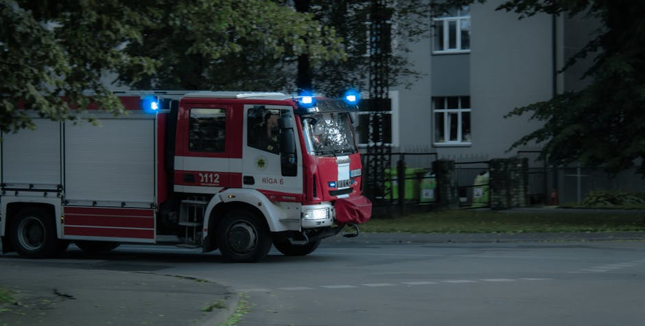 A fire engine parked on a residential street in White City during the daytime, with blue lights flashing on top. The vehicle is red with white and grey details, displaying the emergency number 112 and the city name Riga 6. The front of the fire truck has a large bumper covered with a red protective cloth, and the side features compartments for firefighting equipment. The street is lined with multi-storey residential buildings with white facades and large windows, some trees and greenery are visible along the pavement, and the scene is well-lit, suggesting good natural daylight. This image captures a typical urban setting where emergency services like Man with Van White City might operate in connection with home relocations or emergency moves, highlighting the importance of timely and efficient emergency vehicle access during urgent relocation or logistical scenarios.