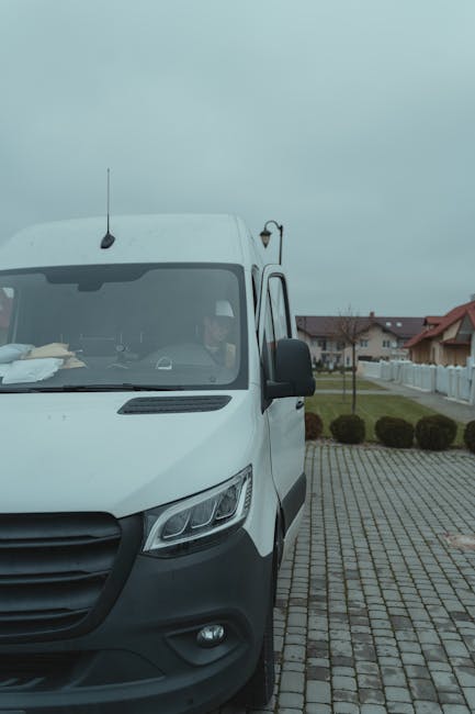 The image shows a white delivery van from Man with Van White City parked on a paved driveway, with the front facing the camera. The van is positioned beside a residential street with a row of houses,