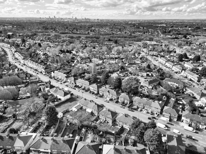 Aerial black and white photograph capturing a residential neighbourhood in White City, showing rows of terraced houses, tree-lined streets, parked cars, and gardens. The streets are busy with cars parked along the curbs, and the houses vary in size with pitched roofs and small front yards. In the foreground, the rooftops of individual homes and backyards are visible, some with fencing and small sheds. Farther in the distance, the urban skyline with taller buildings is faintly visible under a partly cloudy sky. The image provides a detailed view of the typical UK suburban area, suitable for contextual understanding of home relocation, furniture transport, or packing processes involved in house removals, as coordinated by Man with Van White City.
