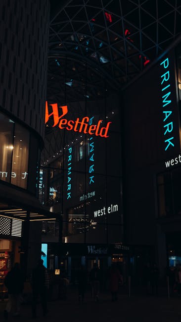 Interior view of a modern commercial building with a high, curved glass ceiling and multiple large, illuminated digital screens displaying the words 'Westfield,' 'Priamark,' and 'West Elm' in bright blue and red text. The space features dark walls, with some areas lit by warm, ambient lighting, and a few figures visible walking or standing inside. This environment is typical of a shopping centre or retail complex, relevant to the context of home relocation and furniture transport, where packing and moving activities might occur within or around such premises. The overall scene emphasizes the scale and design of a shopping mall, supporting the theme of house removals and unloading process in a busy retail setting, as part of a comprehensive move-day checklist at Westfield White City.