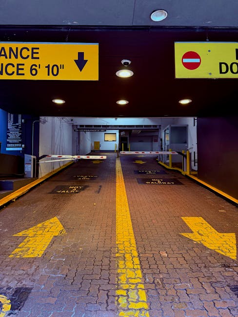 Image of a parking garage entrance with a height clearance barrier overhead displaying a yellow sign indicating a 6 feet 10 inches clearance. The area features a brick-paved driveway with yellow directional arrows and lines guiding vehicles forward, leading into an indoor loading zone. Barriers and bollards are positioned on either side of the entrance to control access. Inside the garage, there are white walls, ceiling-mounted lights, and visible structural elements. This setting is suitable for vehicle access during home relocations and furniture transport, with the company Man with Van White City providing removals services. The overall scene depicts a secure, organized environment for loading furniture and boxes from a property in the process of a house move, emphasizing efficient logistics and proper access planning for moving activities.