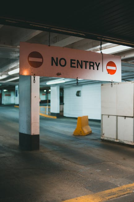 A parking garage with concrete columns and low ceilings, featuring a large rectangular 'No Entry' sign hanging from the ceiling. The sign has a white background with bold black text and red circular no-entry symbols on each side. The garage floor is marked with yellow lines for parking bays, and there are various parking aids such as a yellow wheel stop and white barriers. The area is lit with artificial overhead lighting, creating a utilitarian environment typical for urban building access. This setting relates to move-in or move-out logistics, where access restrictions are common during home relocations managed by companies like Man with Van White City, which specializes in house removals and furniture transport.