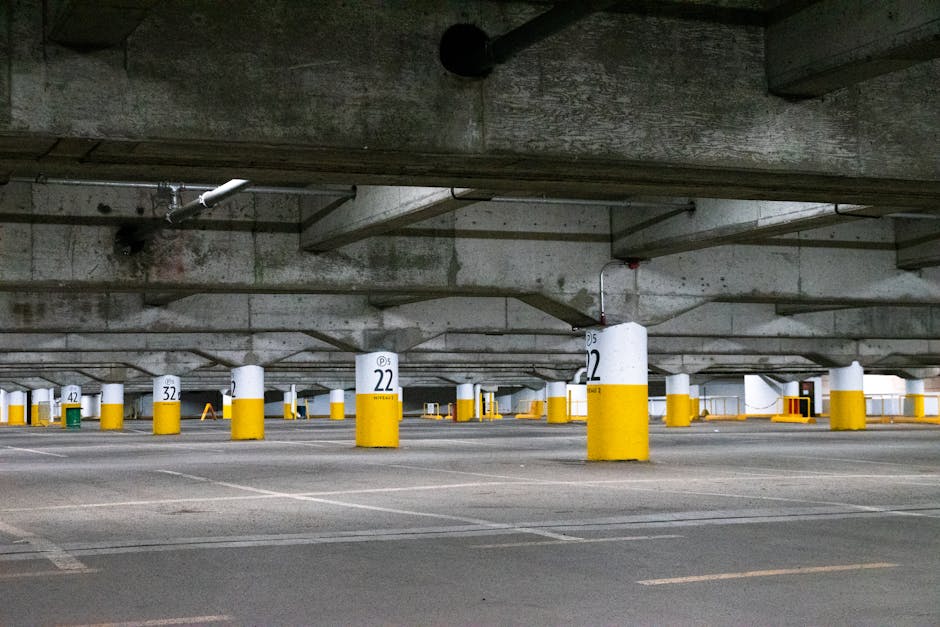 Image of a parking garage entrance with a height clearance barrier overhead displaying a yellow sign indicating a 6 feet 10 inches clearance. The area features a brick-paved driveway with yellow directional arrows and lines guiding vehicles forward, leading into an indoor loading zone. Barriers and bollards are positioned on either side of the entrance to control access. Inside the garage, there are white walls, ceiling-mounted lights, and visible structural elements. This setting is suitable for vehicle access during home relocations and furniture transport, with the company Man with Van White City providing removals services. The overall scene depicts a secure, organized environment for loading furniture and boxes from a property in the process of a house move, emphasizing efficient logistics and proper access planning for moving activities.
