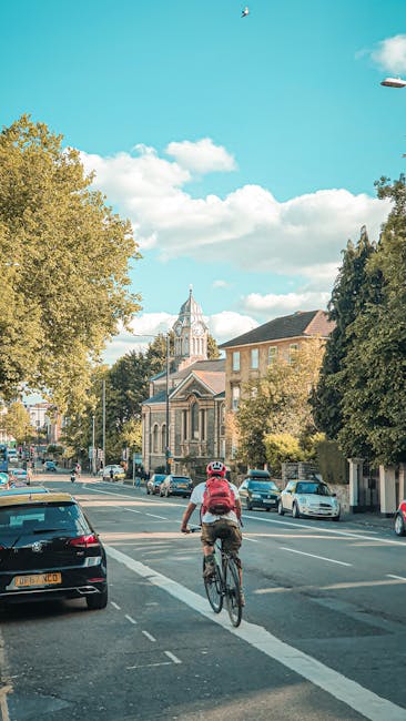A cyclist wearing a pink backpack and a white helmet rides a bicycle along a street lined with parked cars and trees on a bright day with a partly cloudy sky. In the background, there is a historic stone church with a tall clock tower, surrounded by residential buildings and additional trees. The scene depicts an urban setting, possibly within White City, as referenced on the page about house moves and furniture transport. The street appears calm, and the image captures the natural environment alongside the infrastructure relevant to home relocation logistics, with vehicles and a bicycle in the foreground supporting the context of moving services offered by Man with Van White City.