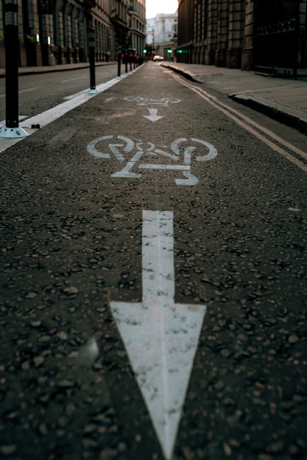 A close-up view of a dedicated cycling lane on a city street, featuring a white painted bike symbol and a downward-pointing arrow on the asphalt surface, which appears slightly textured and worn. The lane is separated from the main road by black metal poles and white painted lines, with the background showing tall urban buildings, some with glass windows reflecting light, and traffic lights at a junction in the distance. The scene is set during daylight with soft natural lighting, and the street appears quiet, suitable for a home relocation or furniture transport process facilitated by professional removal services such as Man with Van White City. The image emphasizes the designated cycle path that may assist in planning moving logistics or transportation routes during house removals in the White City area.