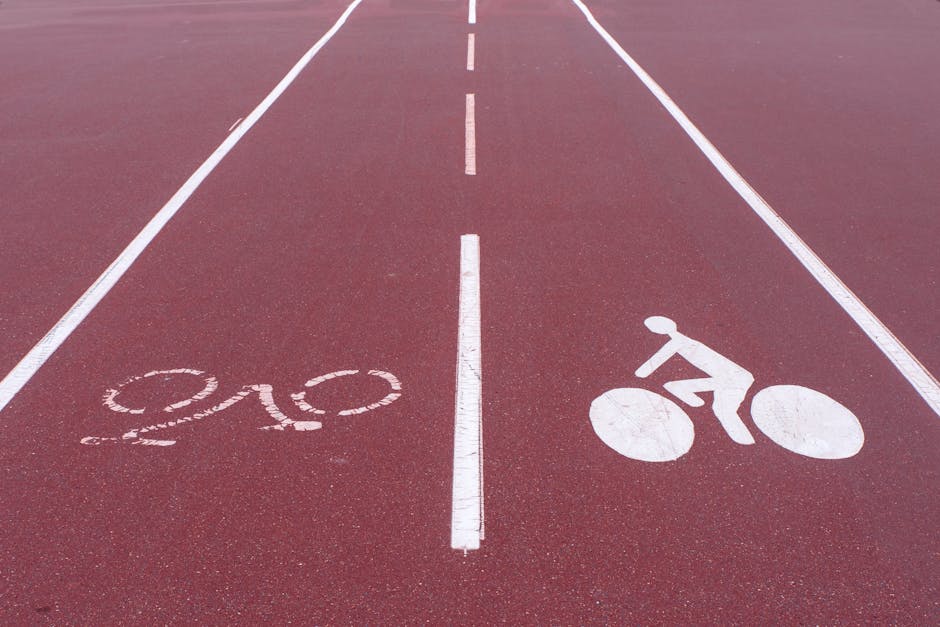 A cyclist wearing a pink backpack and a white helmet rides a bicycle along a street lined with parked cars and trees on a bright day with a partly cloudy sky. In the background, there is a historic stone church with a tall clock tower, surrounded by residential buildings and additional trees. The scene depicts an urban setting, possibly within White City, as referenced on the page about house moves and furniture transport. The street appears calm, and the image captures the natural environment alongside the infrastructure relevant to home relocation logistics, with vehicles and a bicycle in the foreground supporting the context of moving services offered by Man with Van White City.