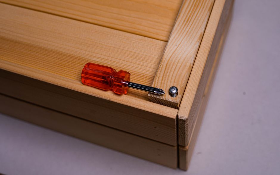 Close-up image showing a flat-pack wooden furniture piece, likely a storage unit or cabinet, with visible light-colored wood panels and assembled corner joints. A red-handled screwdriver is placed on